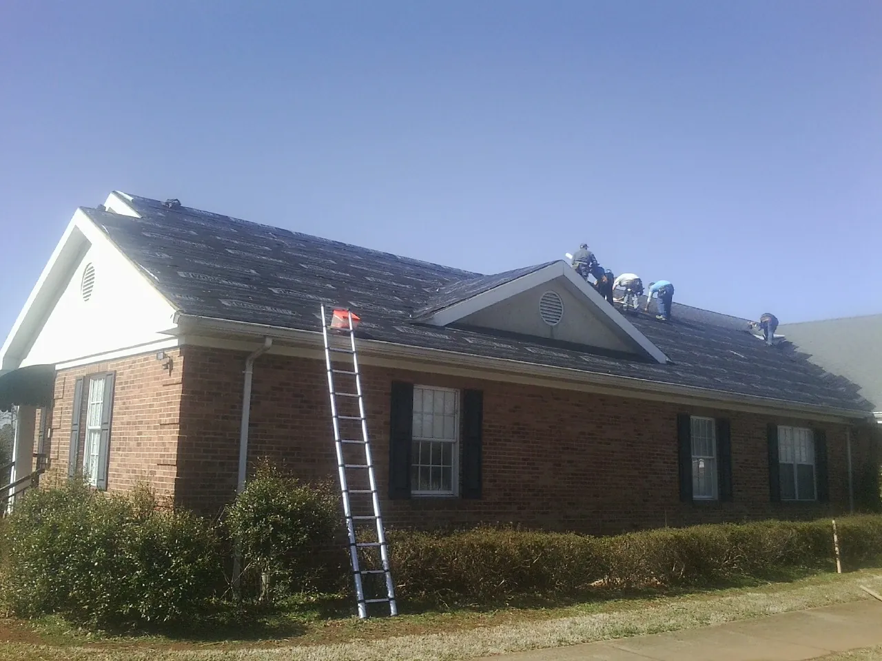 Crew tearing shingles off a brick ranch roof with a ladder set — Metal Roof Repair work by our crew in Walled Lake