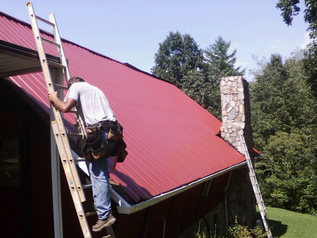 Roofer on a ladder beside a red metal roof and stone chimney — Soffit & Fascia Repair by Roofing Brothers in Walled Lake