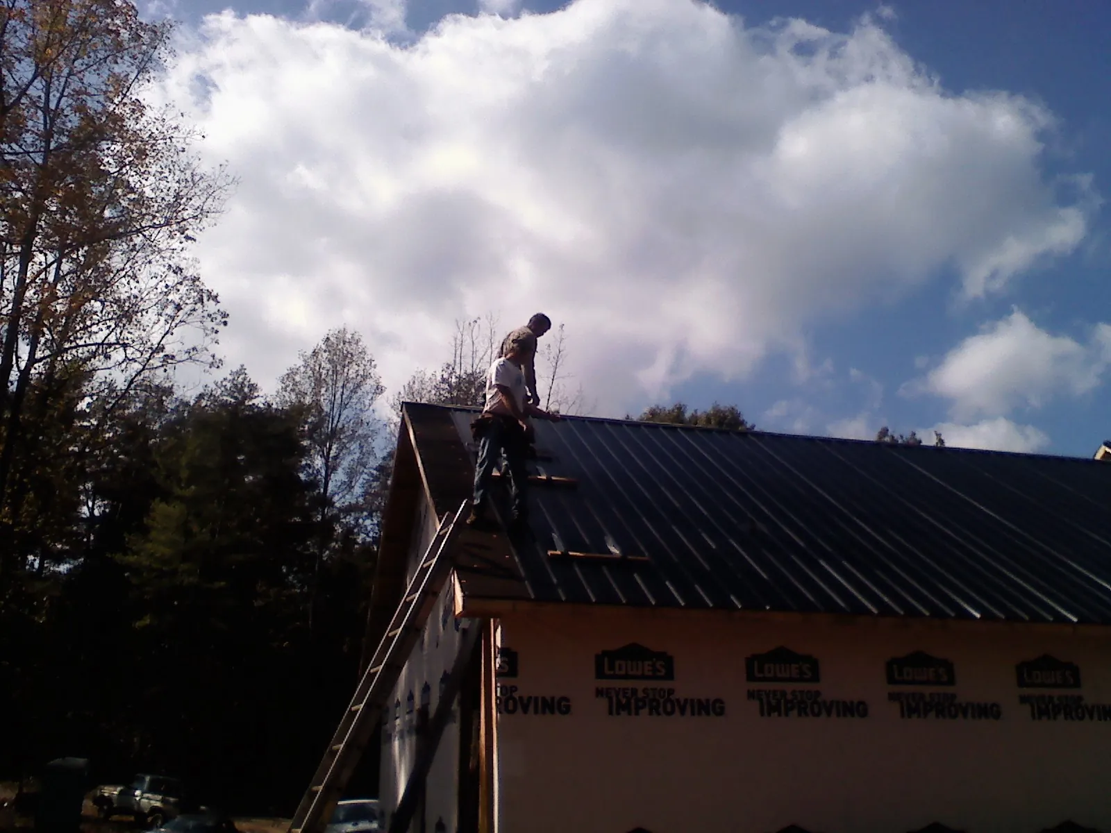 Roofer fastening dark metal panels high against a cloudy sky, from a Roof Inspection job in Walled Lake
